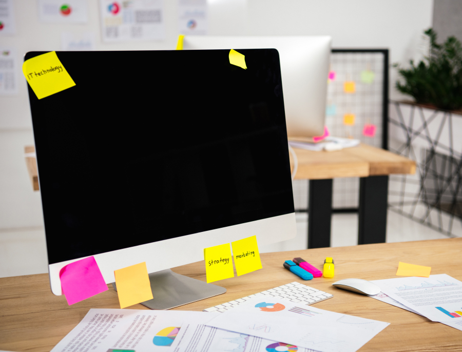 A desktop monitor covered in handwritten sticky notes, with printed charts spread across the desk in front of it