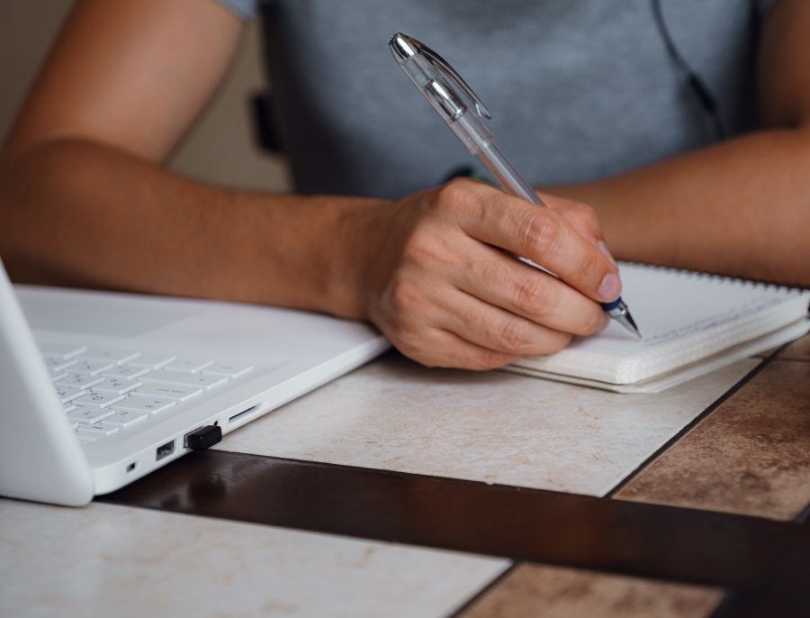 A person writing by hand in a spiral notebook at a desk, with a laptop pushed to one side