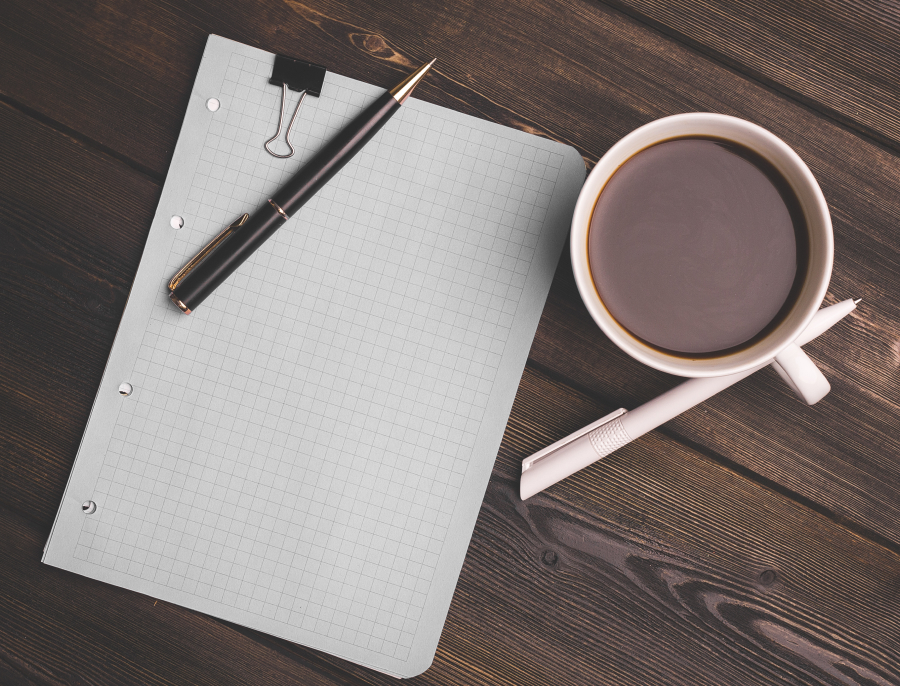 An empty sheet of squared paper with two pens and a cup of coffee on a dark wooden desk, seen from above