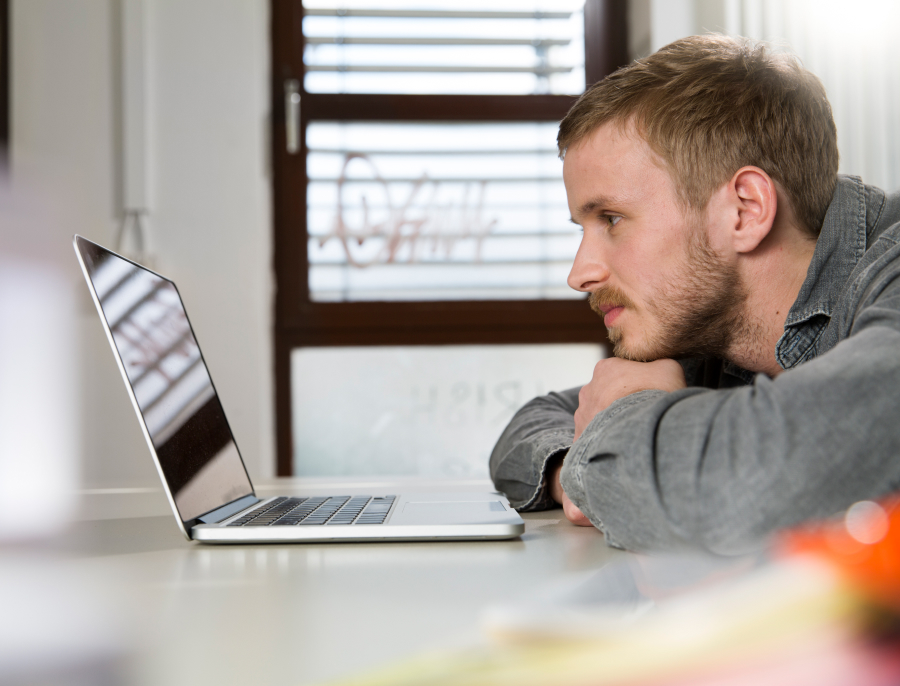 A man resting his chin on his folded arms, looking closely at a laptop screen with a doubtful expression