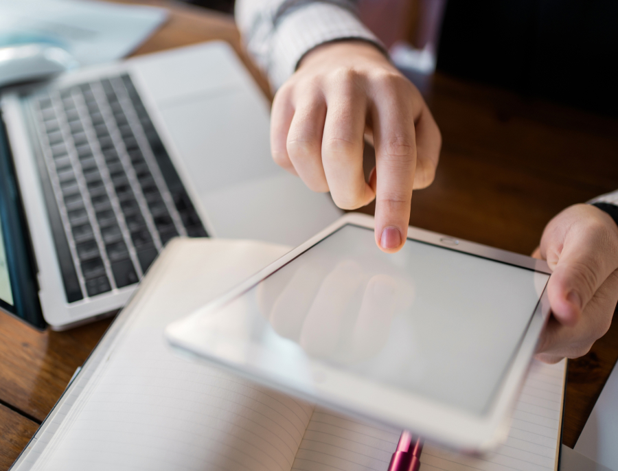 A hand pointing at a tablet screen resting on an open notebook, with a laptop alongside on a wooden desk