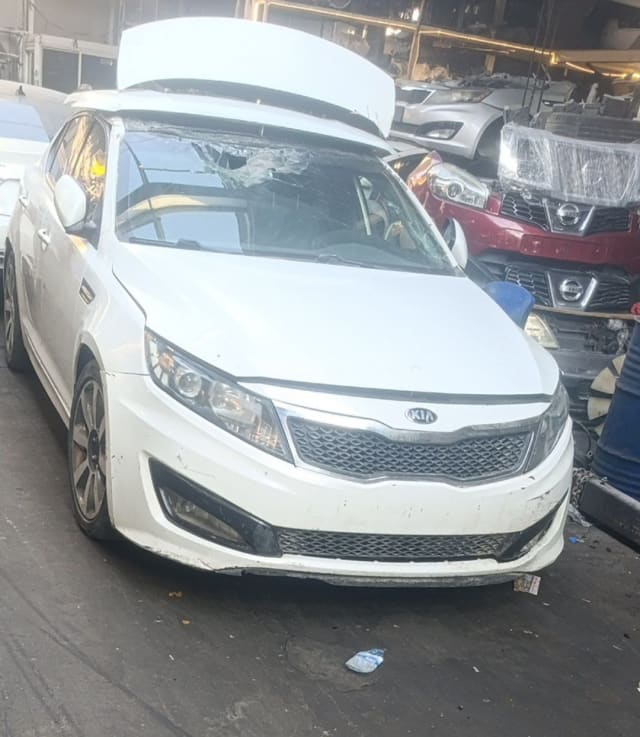 White Kia Optima sedan front — bumper, grille, headlights and raised hood, damaged windshield, Sharjah market