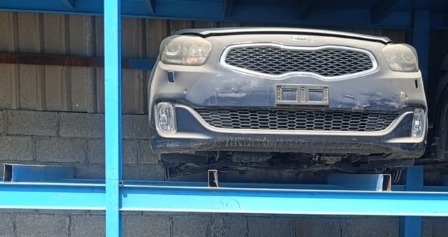 Dark grey Kia Carens front halfcut — bumper, grille, headlights and fog lights on a rack, Sharjah market