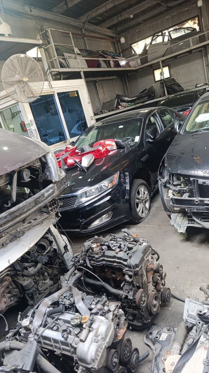 Black Kia Optima sedan for parts — front bumper, grille, headlights and hood, with loose engines nearby, Sharjah market