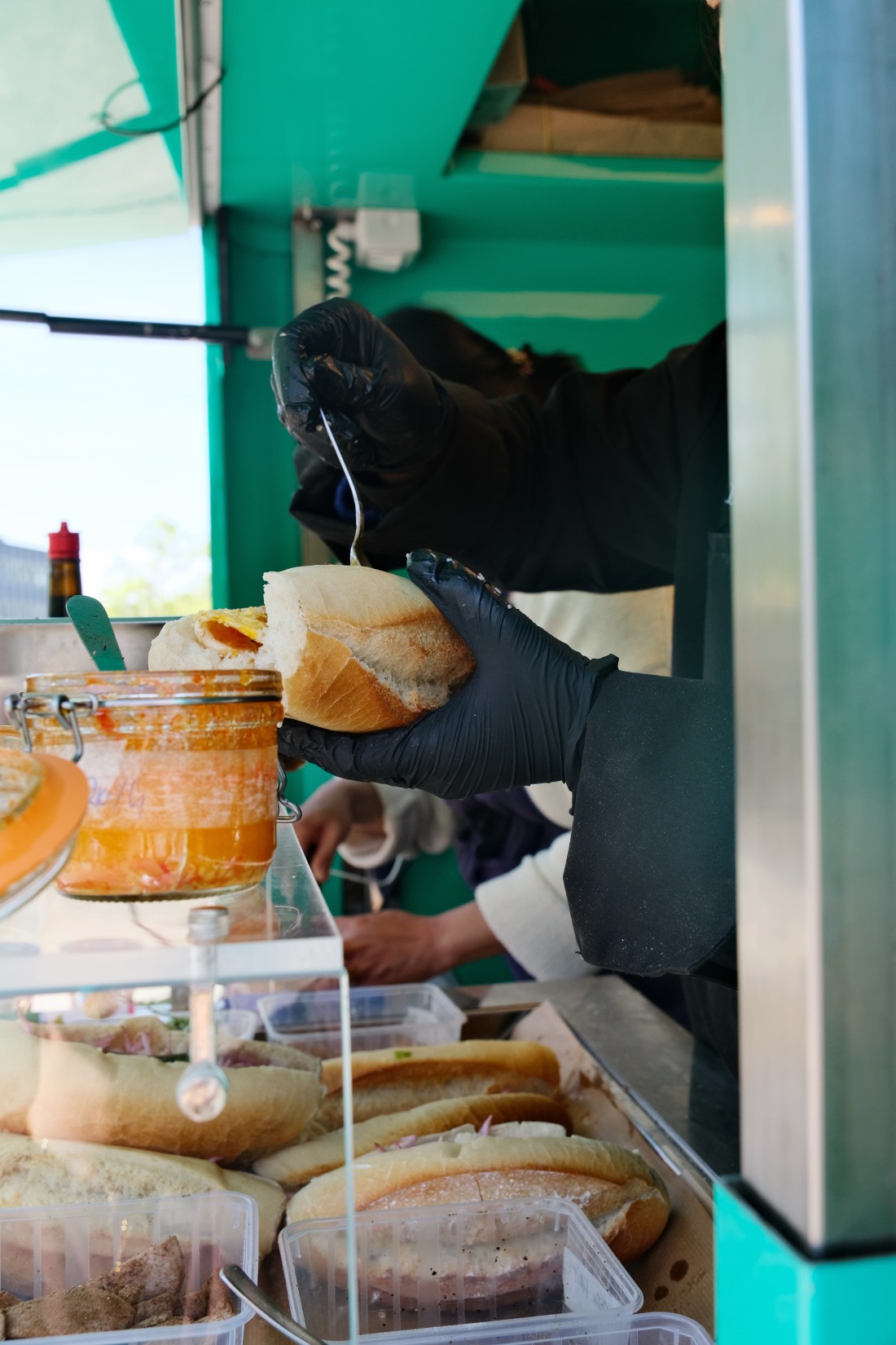 Préparation d'un banh mi en cuisine chez Banh Banh, main gantée noire, baguette et bocaux de condiments