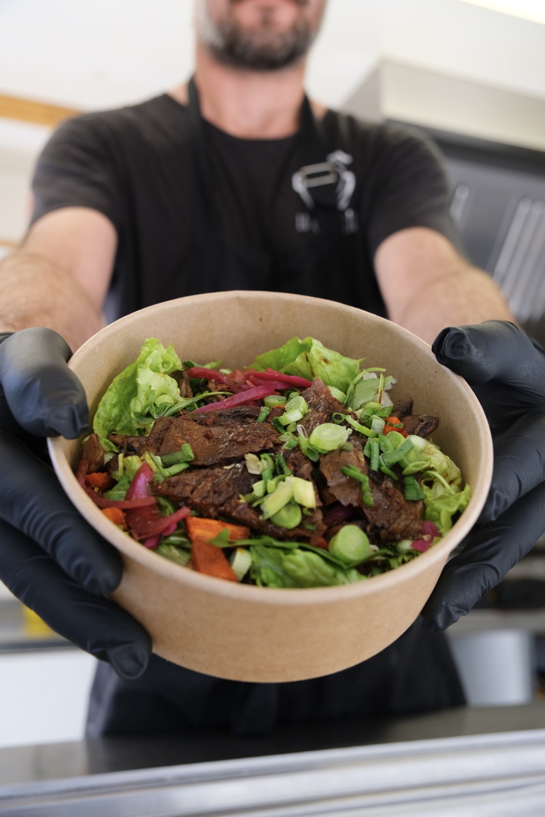 Un bowl signature de La Gamelle servi à deux mains gantées : viande, légumes frais et oignons rouges dans un bol kraft