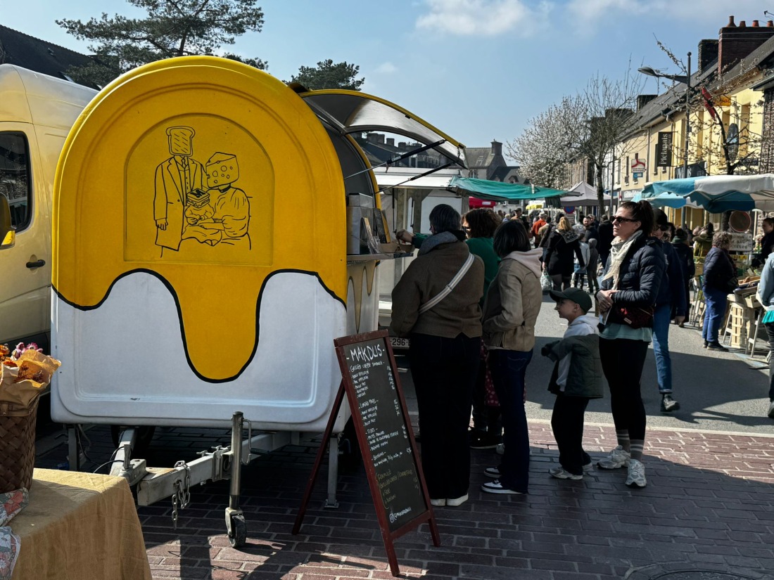 Le food truck Makdus jaune et blanc sur un marché rennais, avec illustration cartoon sur le côté et foule devant le comptoir