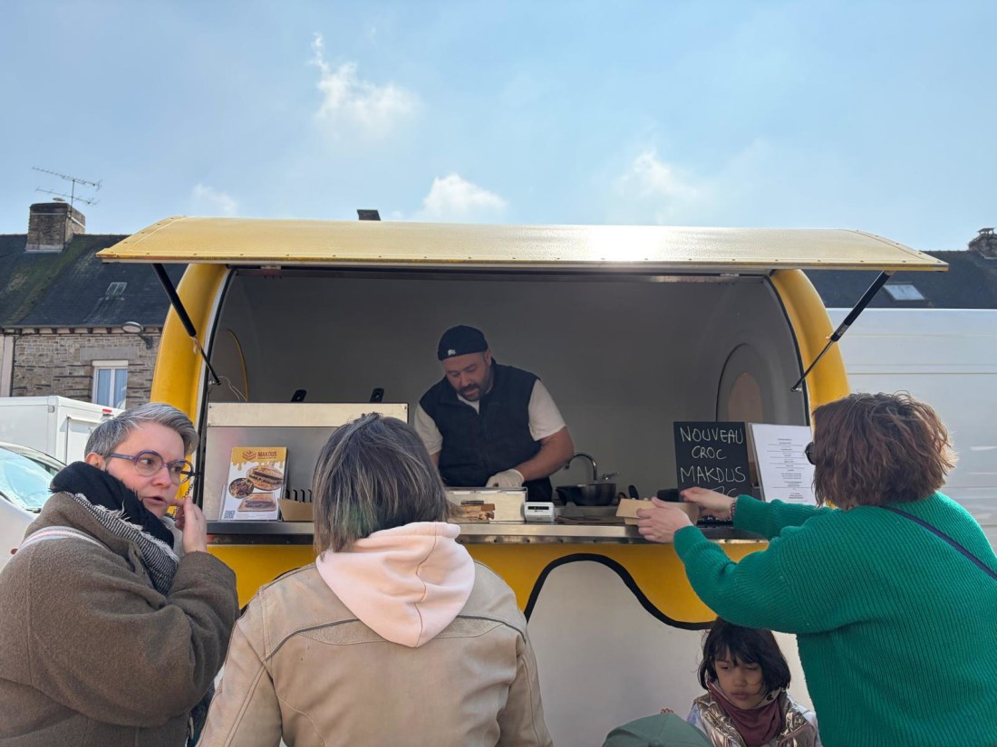 Le chef Makdus en service derrière son comptoir, ardoise « NOUVEAU CROC MAKDUS » visible, trois clientes devant en train de commander