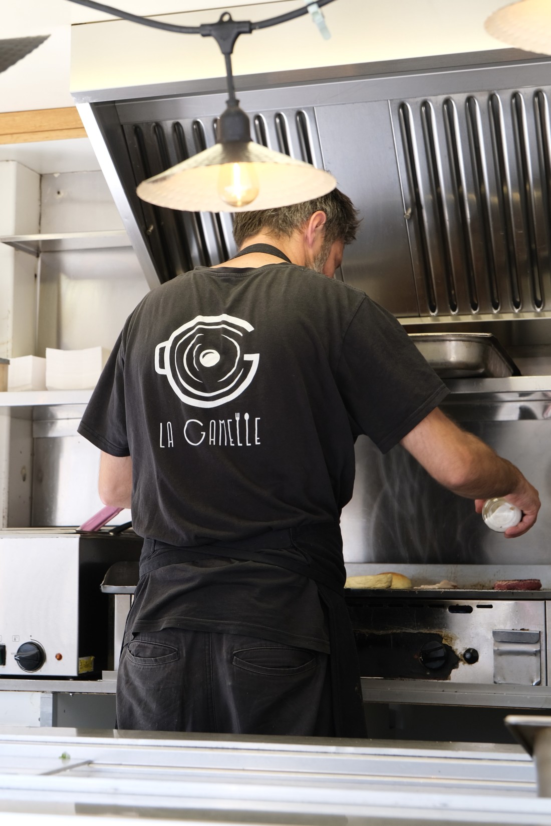 Un cuisinier de dos, t-shirt noir avec le logo de La Gamelle en blanc (marmite stylisée), en train de cuire à la plancha dans le camion