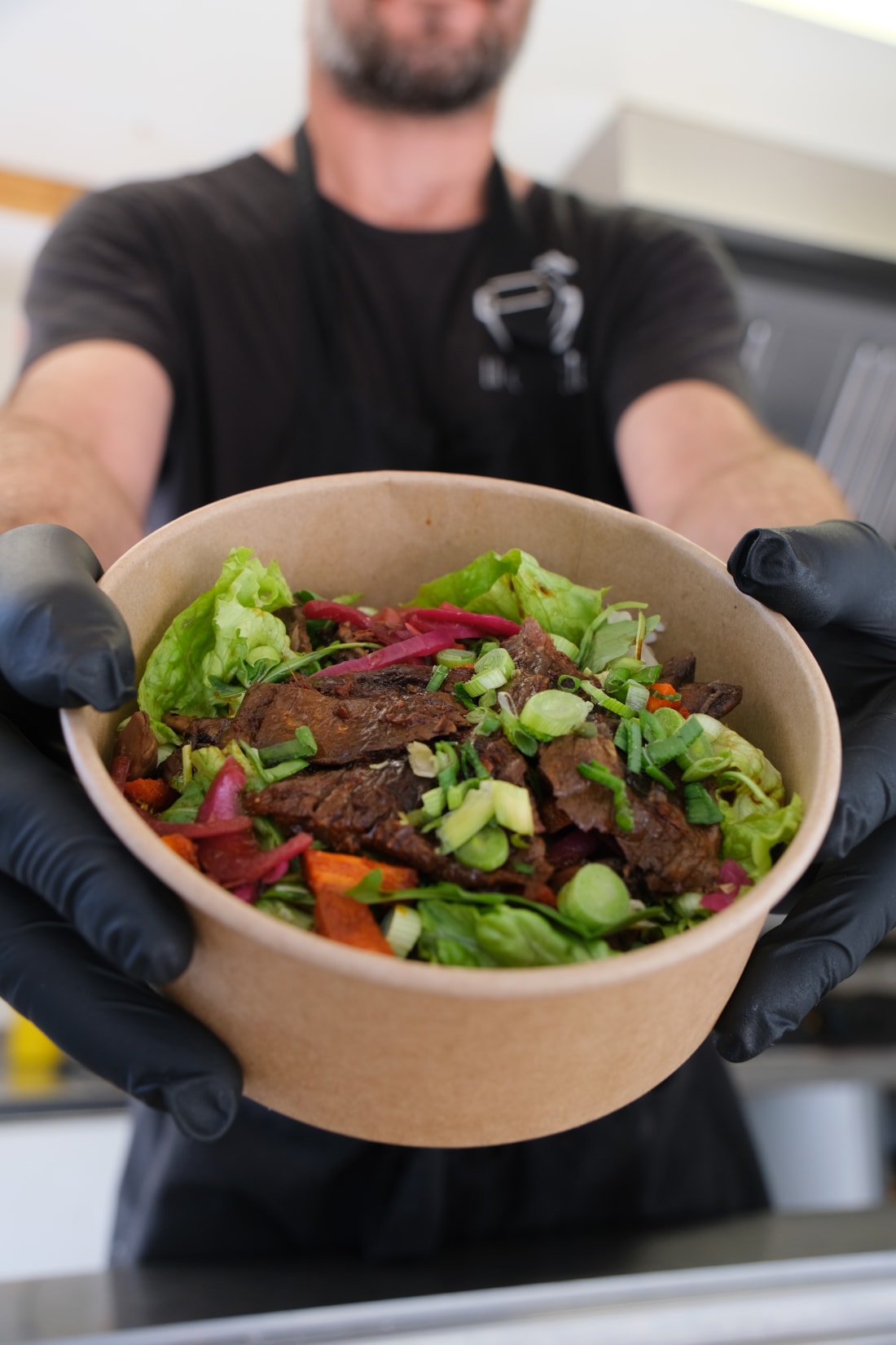 Un bowl steak frais à 2 mains gantées : steak grillé, légumes frais, oignons rouges marinés, vert frais — dressage minute