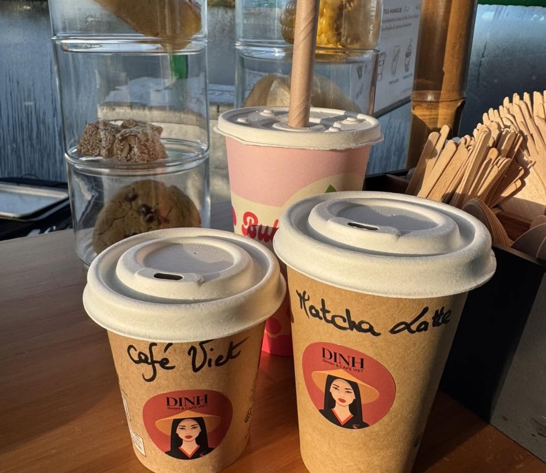 Trois boissons DINH sur un comptoir en bois : un gobelet « Café Viet », un « Matcha Latte » et un bubble tea rose, au logo de la femme au chapeau conique