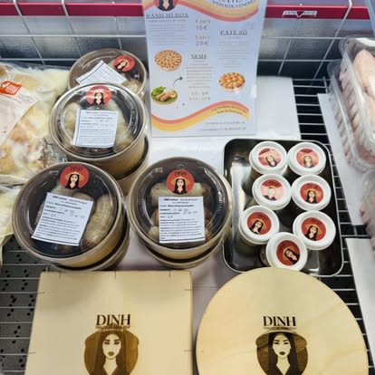 Des produits DINH en présentation : bánh mì box, pots de pâté et de sauces étiquetés au logo DINH, couvercles en bois gravés, dans une vitrine réfrigérée