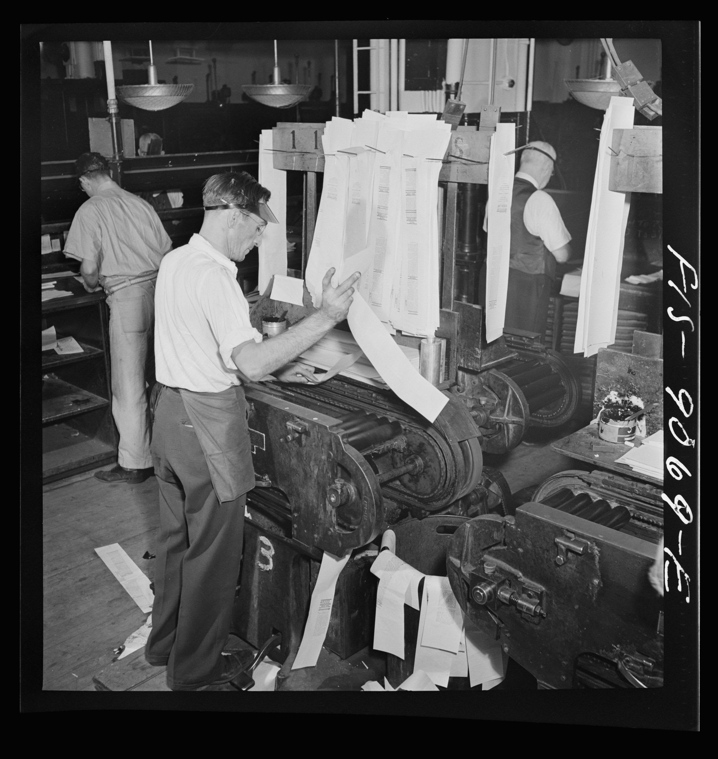 A pressman in an eyeshade and apron feeds a proof press in a newspaper composing room. Long strips of galley proofs hang from a rack above the press and spill onto the floor beneath it. The whole negative is shown, inside its black border, with the negative number FS-9069-E handwritten along the right edge.