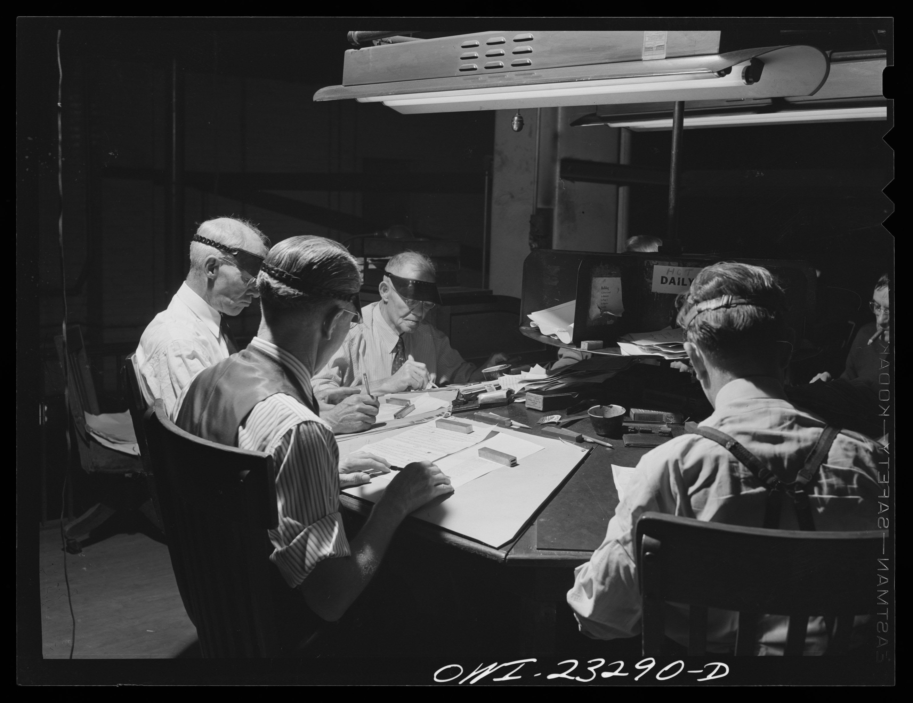 Proofreaders in eyeshades at a newspaper’s proofreading desk, under a hanging fluorescent lamp. Two older men mark sheets with pens; a man in a striped shirt in the foreground reads a sheet held down by metal weights, and a younger man in suspenders reads at the right. The whole negative is shown, inside its black border, with the negative number OWI-23290-D handwritten along the foot.