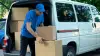 Delivery man in a blue uniform and cap loading brown boxes into a white van