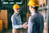 Two gentlemen wearing hard hats are shaking hands in a warehouse