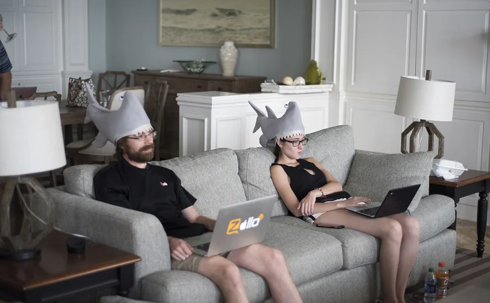 Mann und Frau sitzen mit ihren Laptops auf einem Sofa. Sie haben Haifischhüte auf dem Kopf.