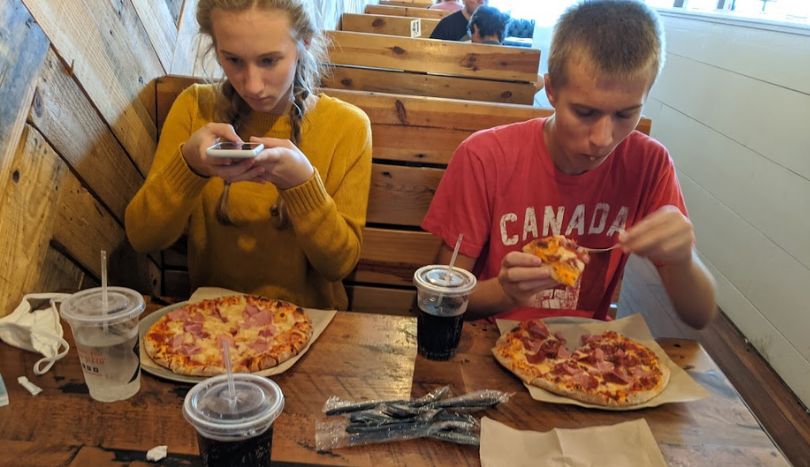 Girl is taking picture of pizza and a boy is eating a pizza