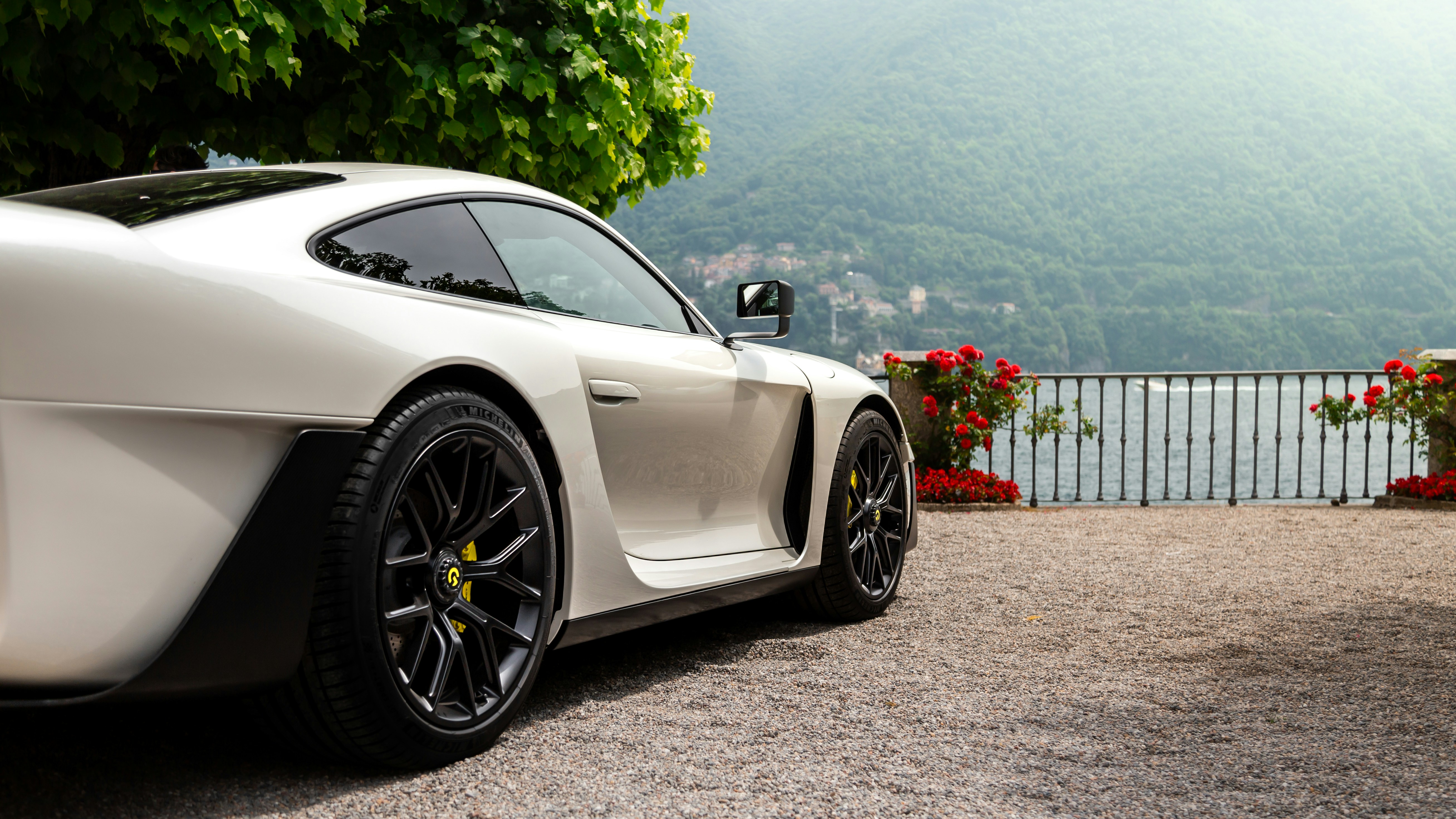 White sports car parked facing a flower-covered fence and beyond it, a lake or ocean flanked by a tall, forested hill.