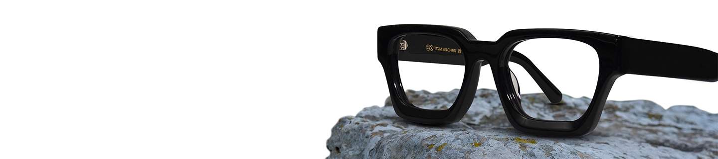 A close up image of black rectangular eyeglasses featuring a chunky black acetate frame placed on a textured stone surface. The image has content that says black glasses and a tagline. It also has a discount offer by Specscart and Trustpilot reviews.