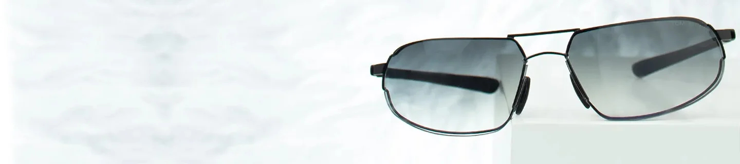 Close-up of sleek brow bar sunglasses with dark-tinted lenses resting on a white geometric block against a soft, minimalist white background.