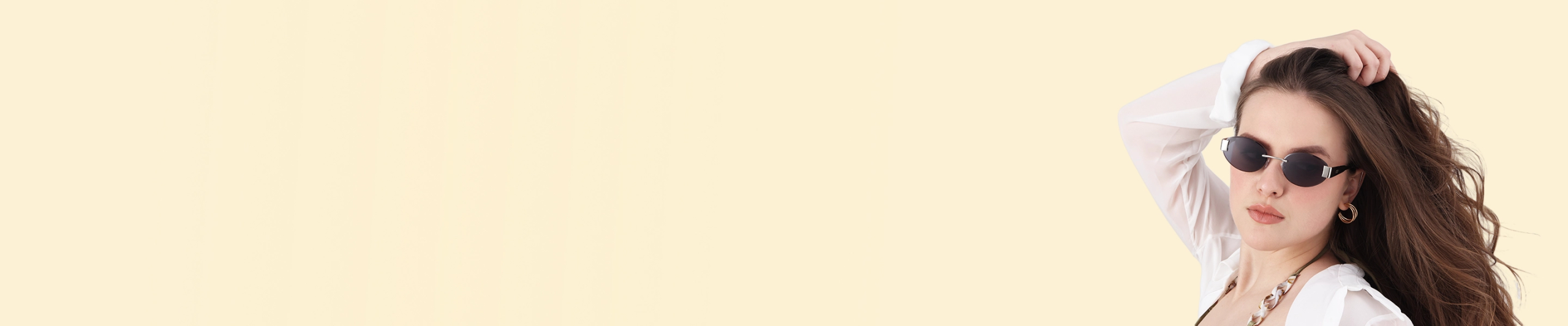 A woman with wavy hair posing in small rimless oval sunglasses with dark lenses and gold hoop earrings against a minimalist light cream backdrop.