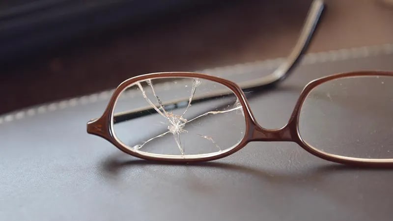 Close-up of brown-rimmed glasses resting on a dark surface, featuring a heavily cracked and shattered left lens with a central impact point.