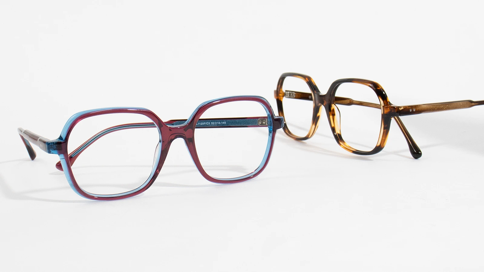 The image has two stylish pair of eyeglasses placed on a greyish-white background. The first pair has an oversized shape with gradient mixing maroon and blue colours. The other pair also has an oversized design but in a tortoiseshell pattern.