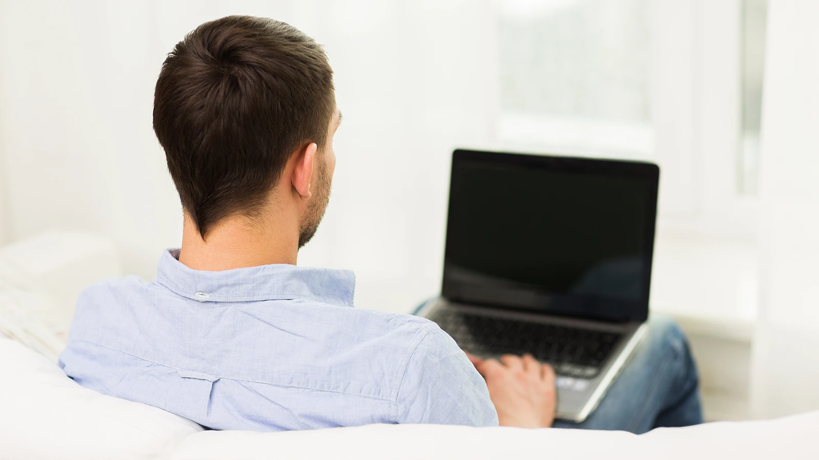 The image features a man sitting on the sofa comfortably while using his laptop that is placed on his legs. It has been taken from behind and has big windows in a blurred background.