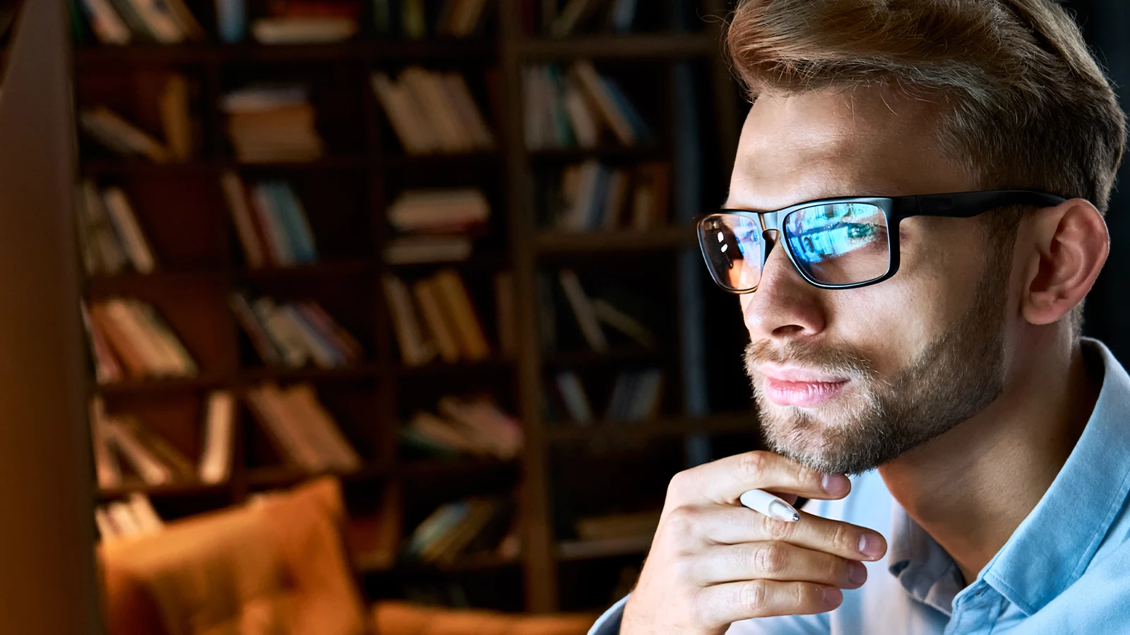 A focused man wearing blue light glasses gazes at a screen, with digital reflections visible in his lenses. The warm library backdrop contrasts with the cool glow, which shows the glasses' protective and stylish appeal.