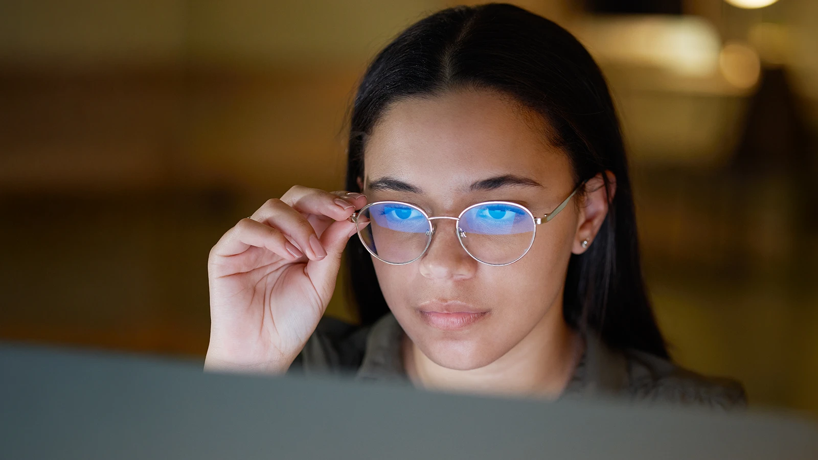 A woman wearing blue light-filtering glasses focuses intently on a screen, adjusting her frames for clarity. The soft glow of the monitor is reflected in her lenses, showing a serious research session.