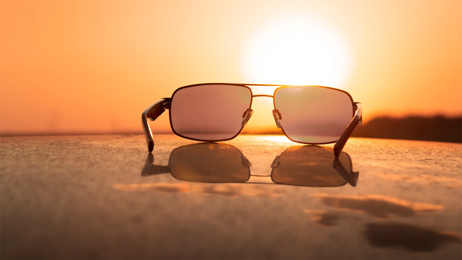 A sleek pair of rectangular sunglasses rests on a reflective surface, capturing the warm glow of a golden sunset. The design highlights the lenses and frame, symbolising style, relaxation, and timeless summer vibes.