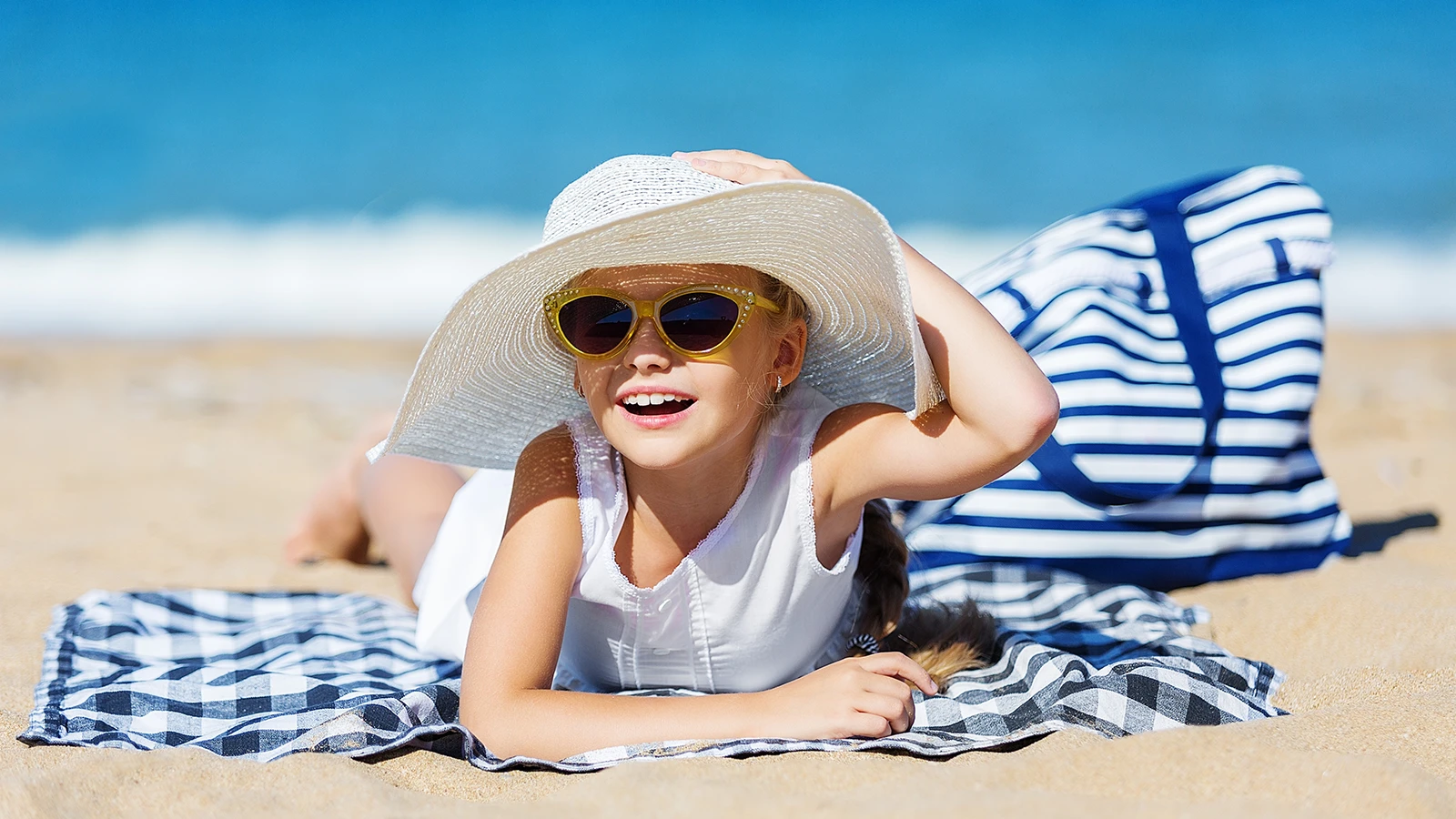 A cheerful young girl relaxes on the beach, wearing stylish yellow cat-eye sunglasses and a wide-brimmed white hat. Dressed in a sleeveless white outfit, she smiles brightly, enjoying the sunny day beside a striped beach bag.