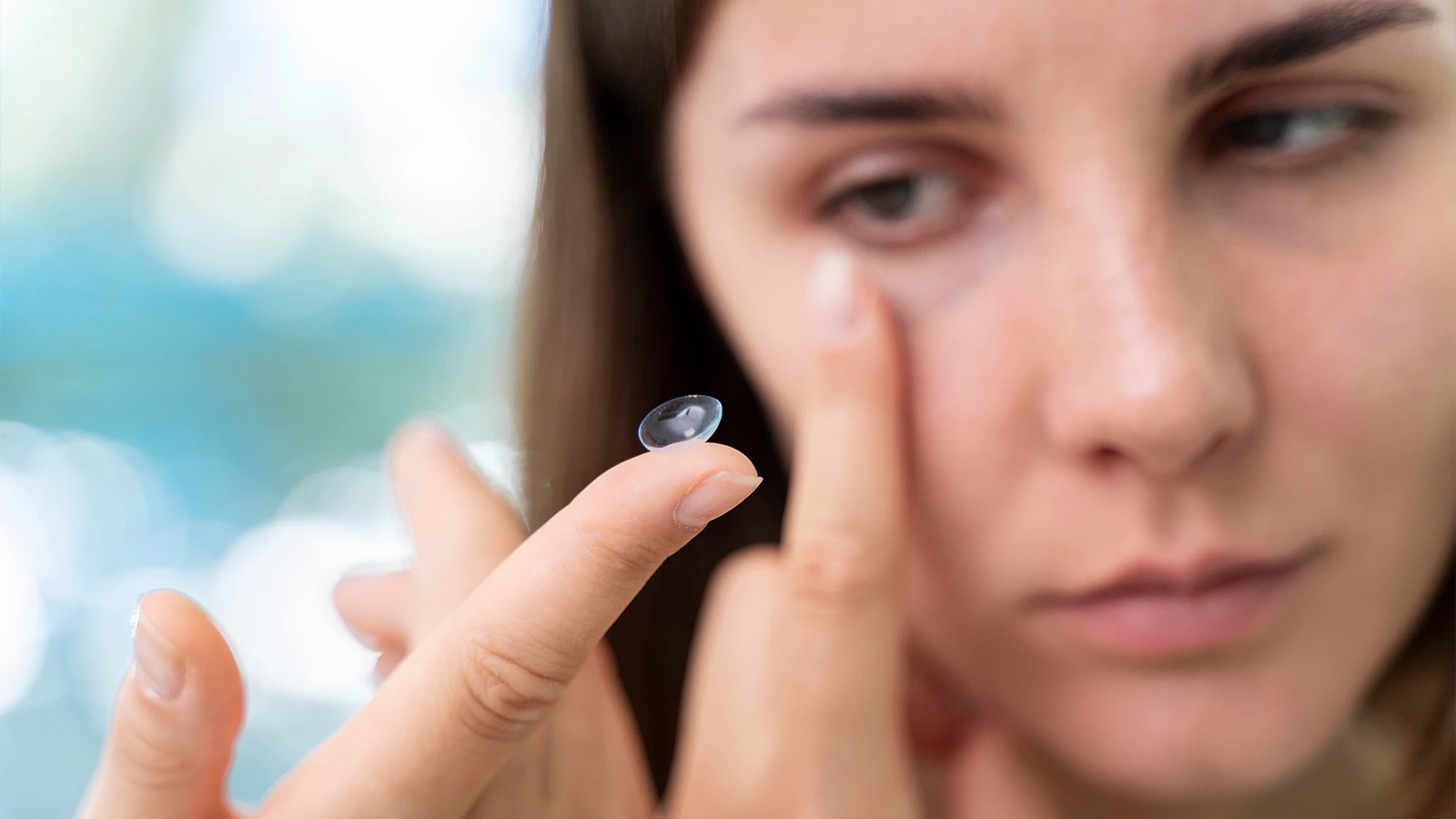 A woman carefully prepares to insert a contact lens into her eye. The lens rests delicately on her fingertip, while her other hand gently pulls down her lower eyelid, reflecting precision and focus in the process.