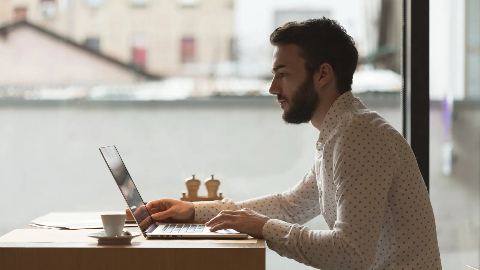 A person wearing a white shirt is sitting at a table and using a laptop. There is a clean setup of office desk a cup and saucer along with papers next to the laptop. A window is visible in the background, showing an out-of-focus urban scene.