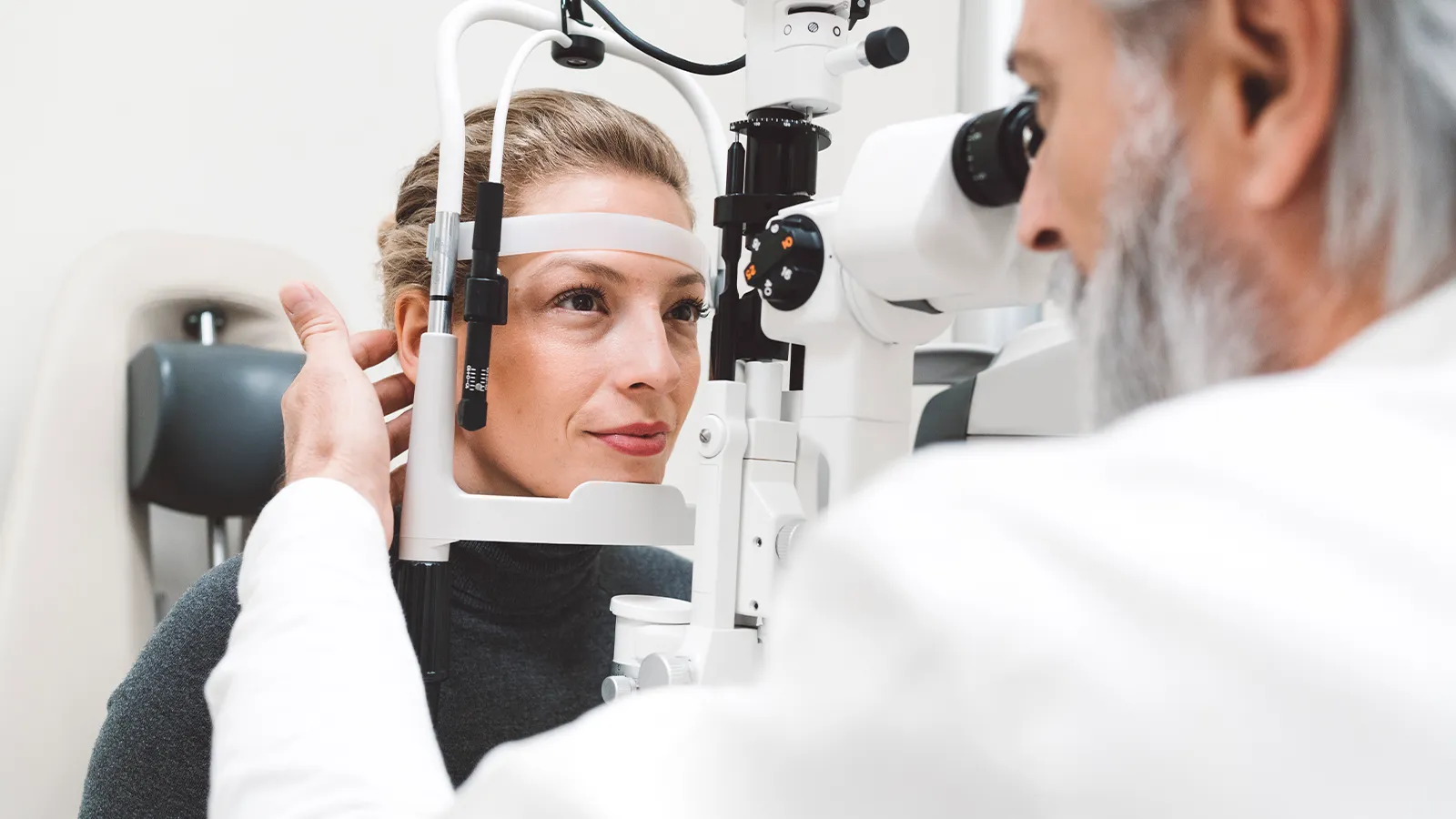A person is having their eyes examined by an eye care professional, who is looking into a specialized ophthalmology machine. The person is holding their head against a support while the professional adjusts the equipment.