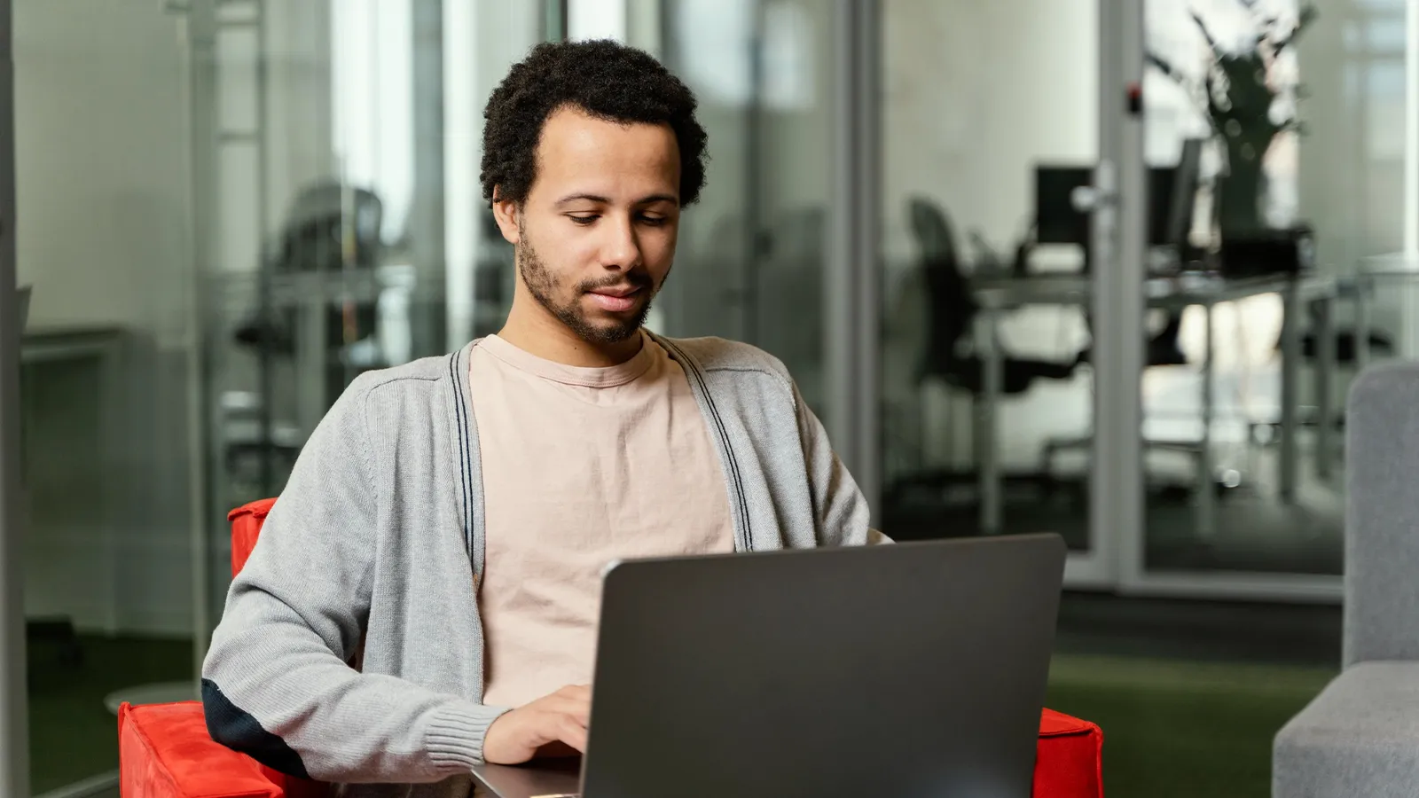 A person with short, dark curly hair and a beard is sitting indoors, focused on a laptop. They are wearing a light-coloured t-shirt and a gray cardigan. The background appears to be an office with glass partitions.
