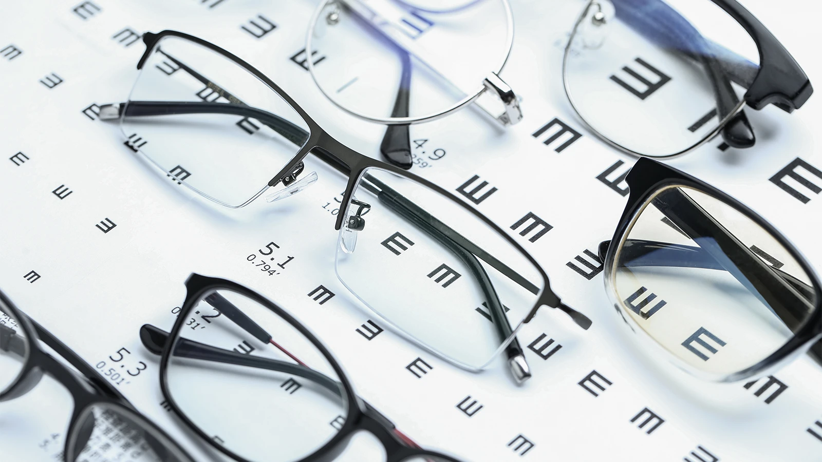 A close-up, high-angle shot of several pairs of eyeglasses scattered on a white background with a large E chart, which is used for vision testing. The glasses have various frame styles, and the chart's letters are visible through the lenses.