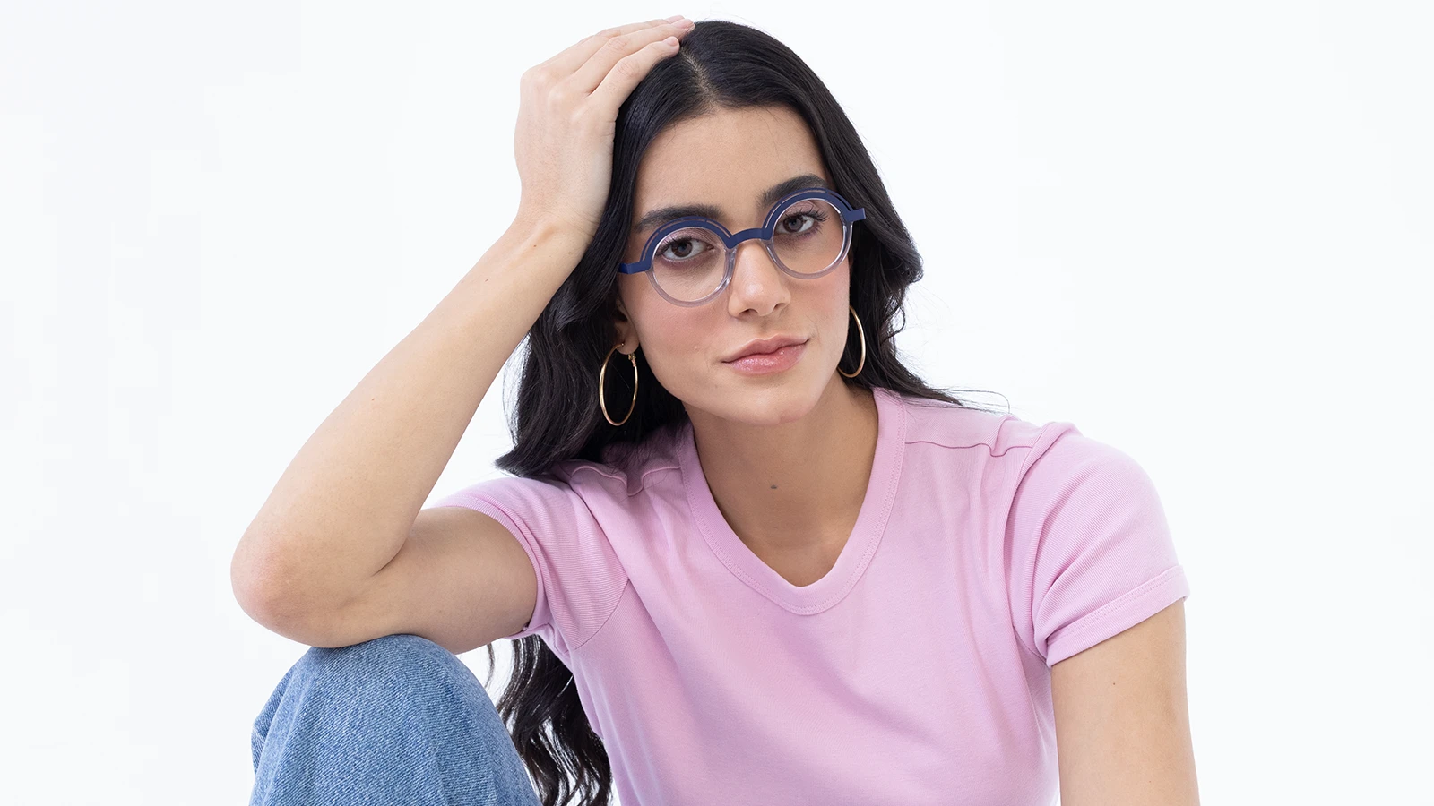 A person with long, dark hair is wearing a pink t-shirt and blue-rimmed glasses. They are seated in front of a white background, resting their head on one hand and looking directly at the camera. They are also wearing large, gold hoop earrings.
