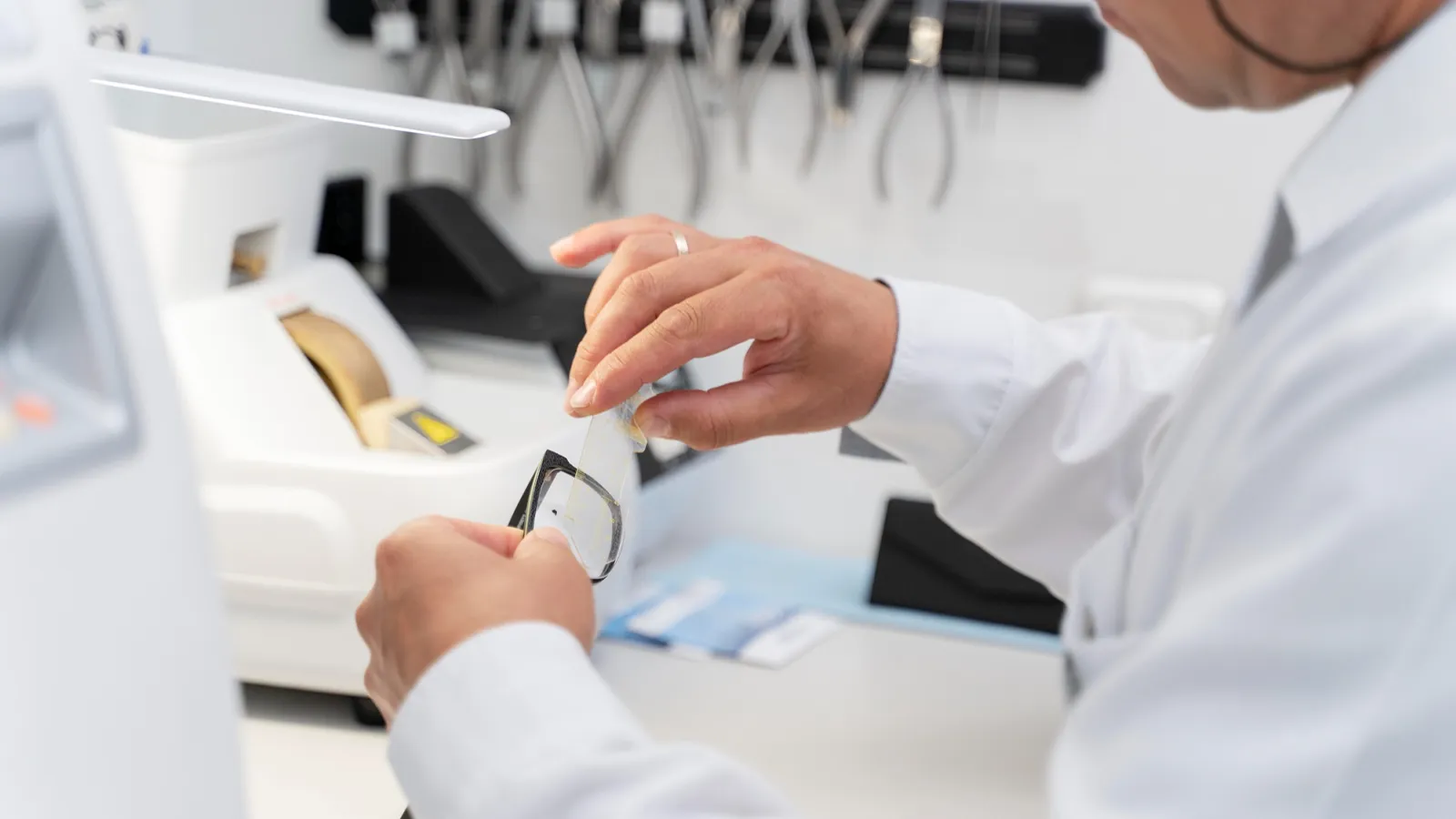 A professional carefully working on a pair of eyeglasses, holding a lens with precision. Surrounded by specialised optical tools, the scene highlights the delicate craftsmanship and accuracy involved in preparing and fitting eyewear.