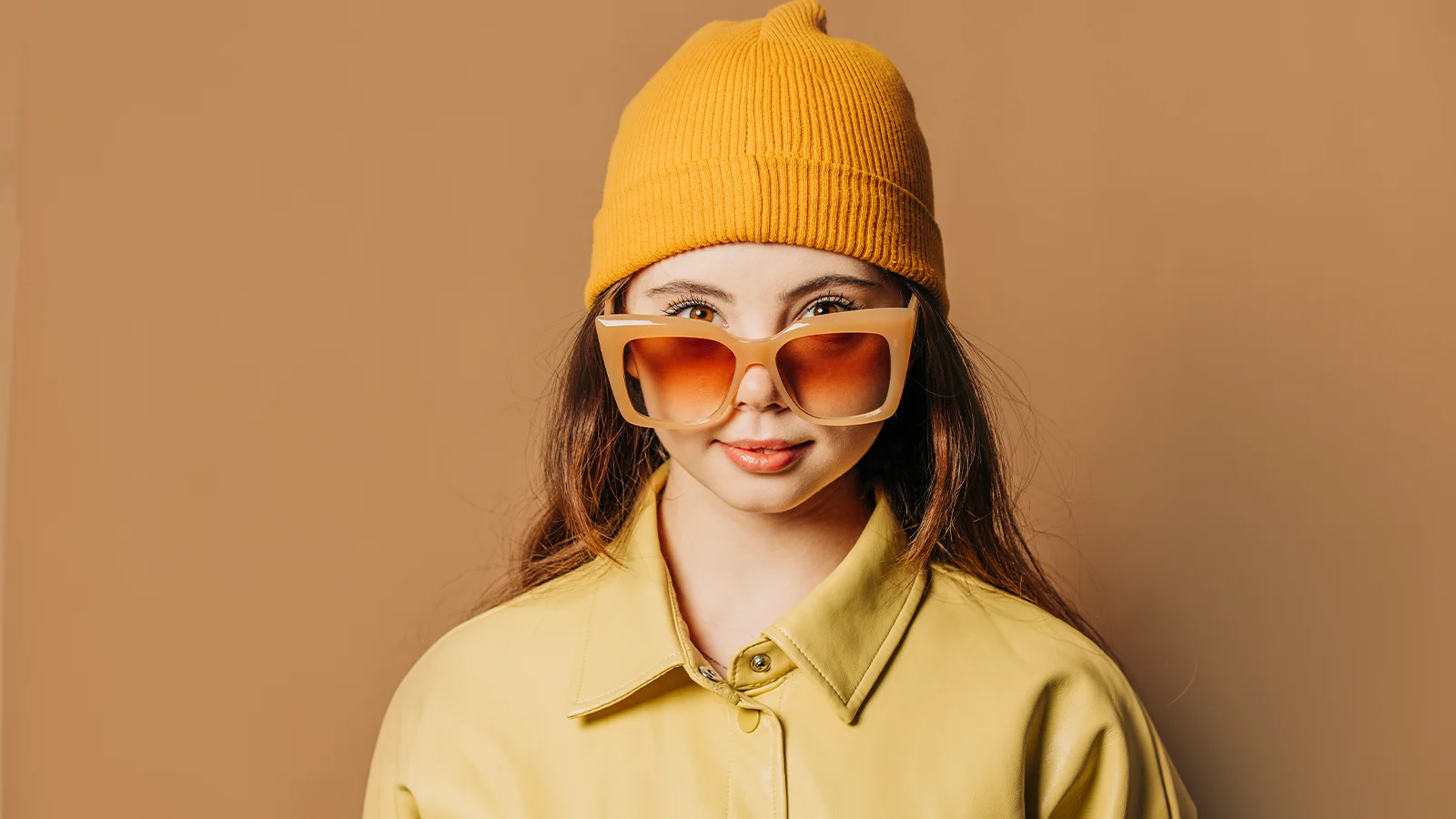 The image has a young woman with open hair wearing a yellow shirt. She is wearing a yellow beanie hat with oversized orange sunglasses.