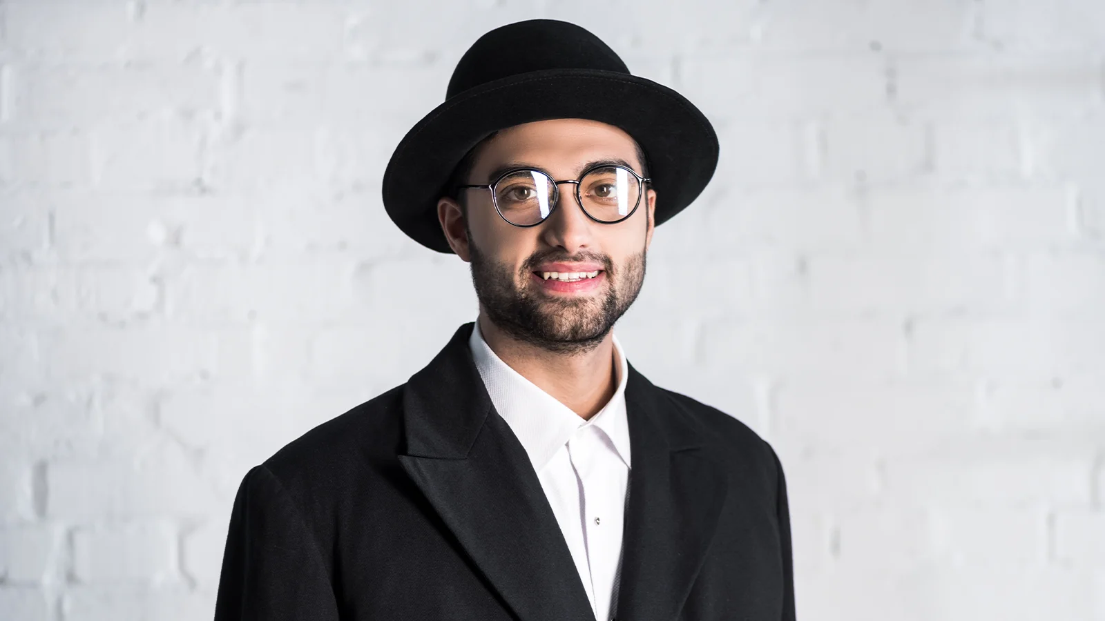 In the image a man is standing wearing a black blazer and white shirt. He has a smile on his face and he is wearing fedora hat with round black metal glasses.
