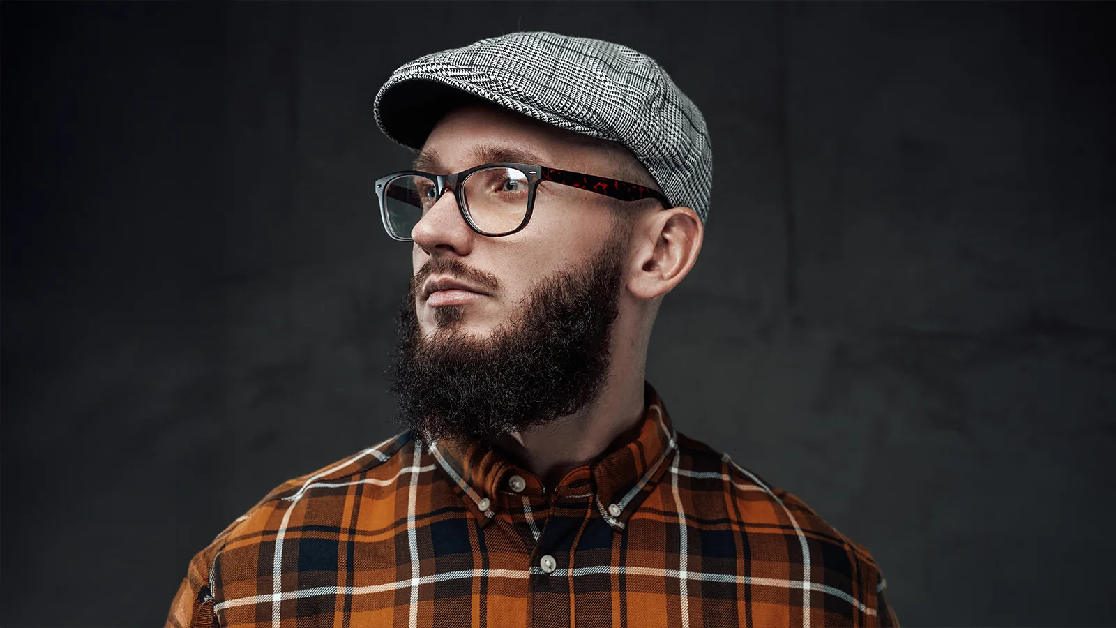 A bearded man is wearing rectangular black eyeglasses with baker boy cap in grey colour with black checkered style. He is also wearing a plaid shirt and he has side facing pose.