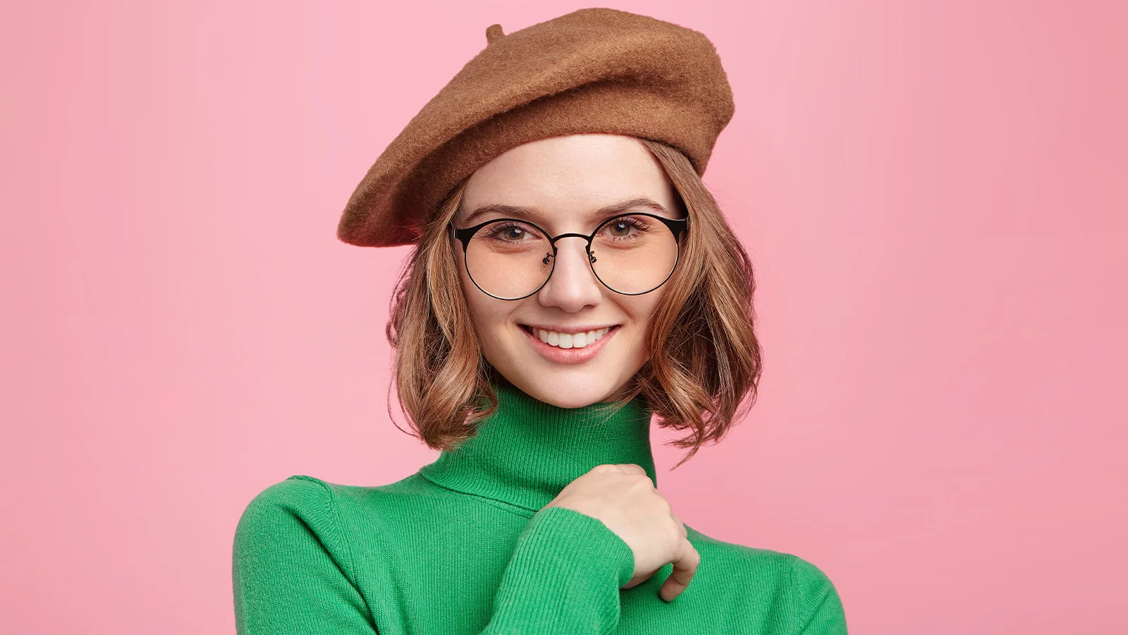 The image has a smiling girl wearing green colour turtleneck ribbed top. Her hand is placed over her shoulder and she is wearing black round glasses and brown beret hat.