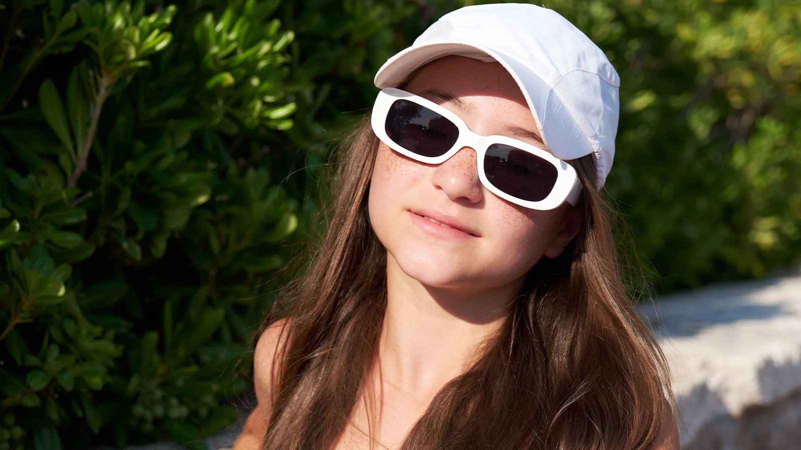 Young woman wearing oversized white rectangular sunglasses and a white cap, with long brown hair, standing outdoors in sunlight against a green leafy background.