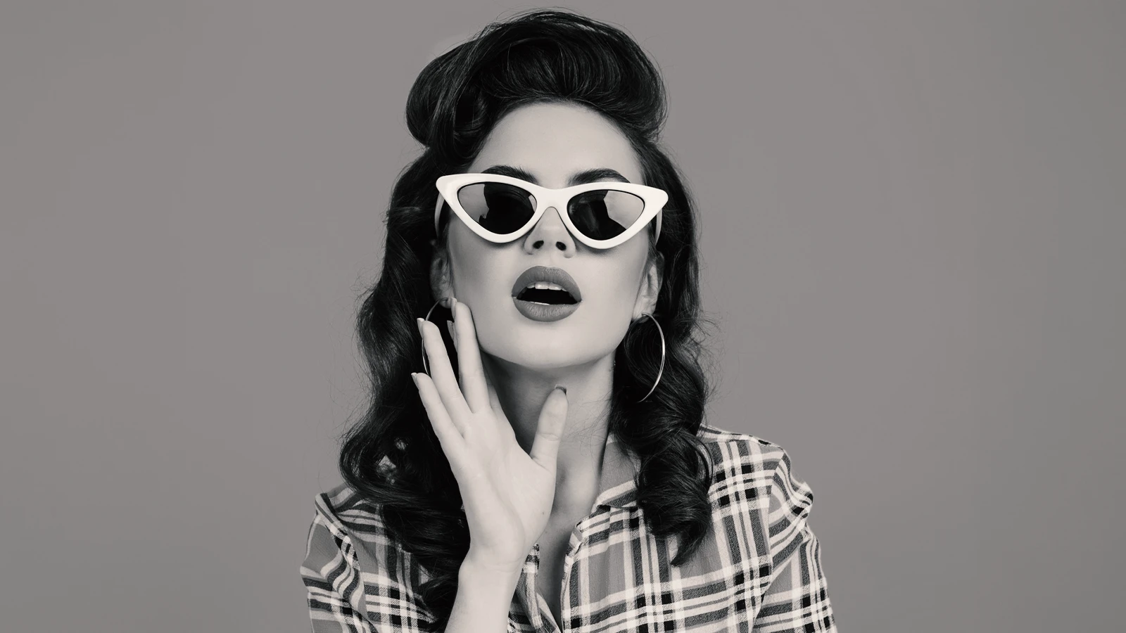 This black and white image captures a woman in a retro 1950s look with voluminous hair and dark lipstick. She is wearing classic, thick-framed white cat-eye sunglasses, an iconic accessory of the era, and a plaid top.