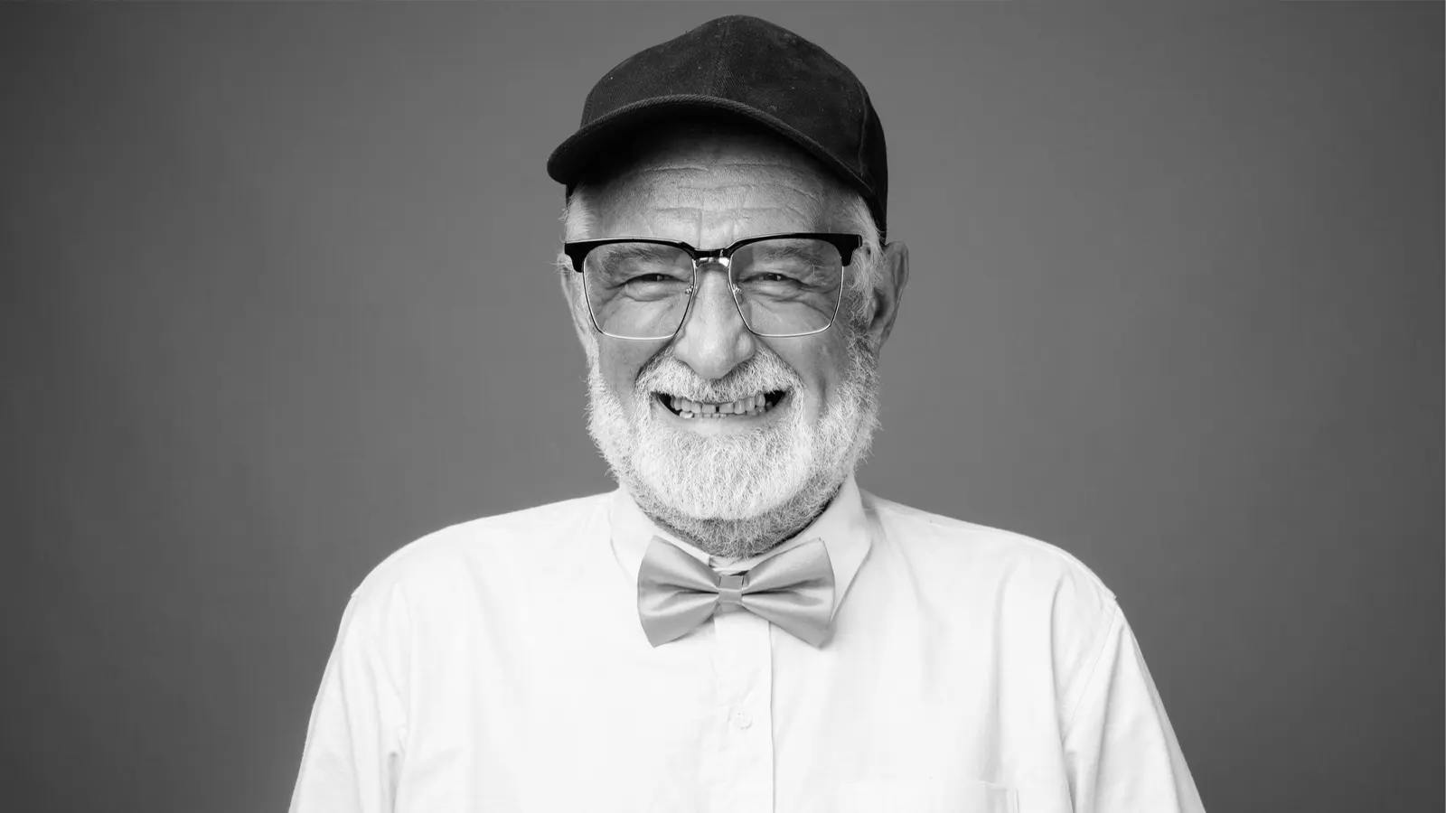 This is a black and white portrait of an older man. He is wearing a dark baseball cap, a white button-down shirt, and a light-colored bow tie. He is smiling widely, and his clear-rimmed eyeglasses with a bold, partially dark frame (often called a 'browline' style) are a significant feature.