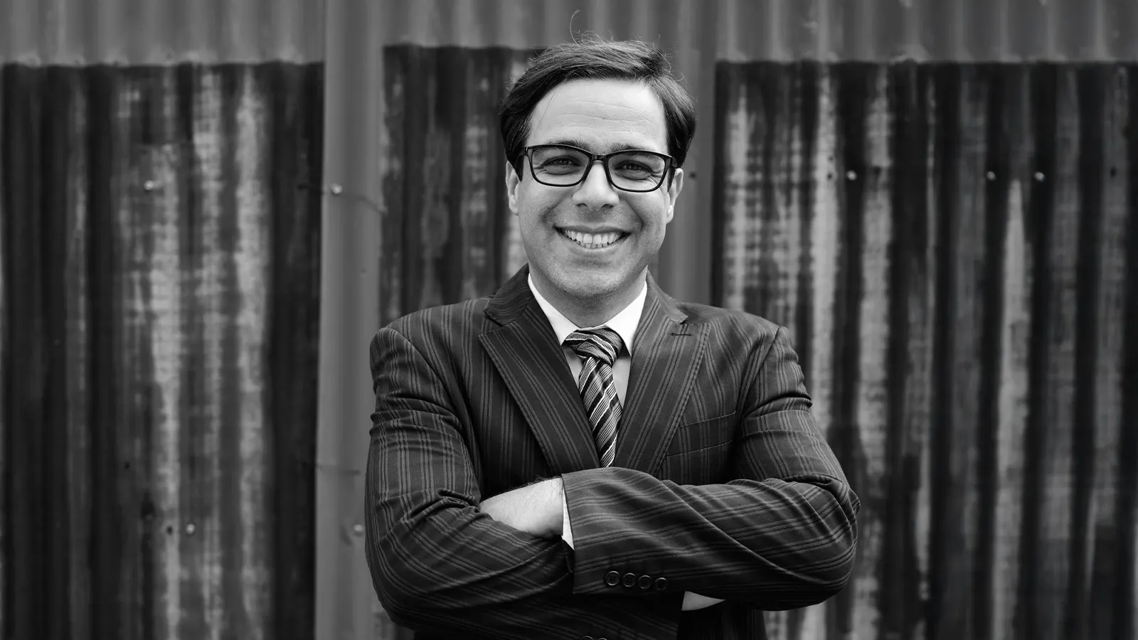 This is a black and white portrait of a man in a business suit, smiling confidently with his arms crossed. He is wearing thick-rimmed, rectangular glasses in a dark colour. These more blocky, square frames became highly popular.