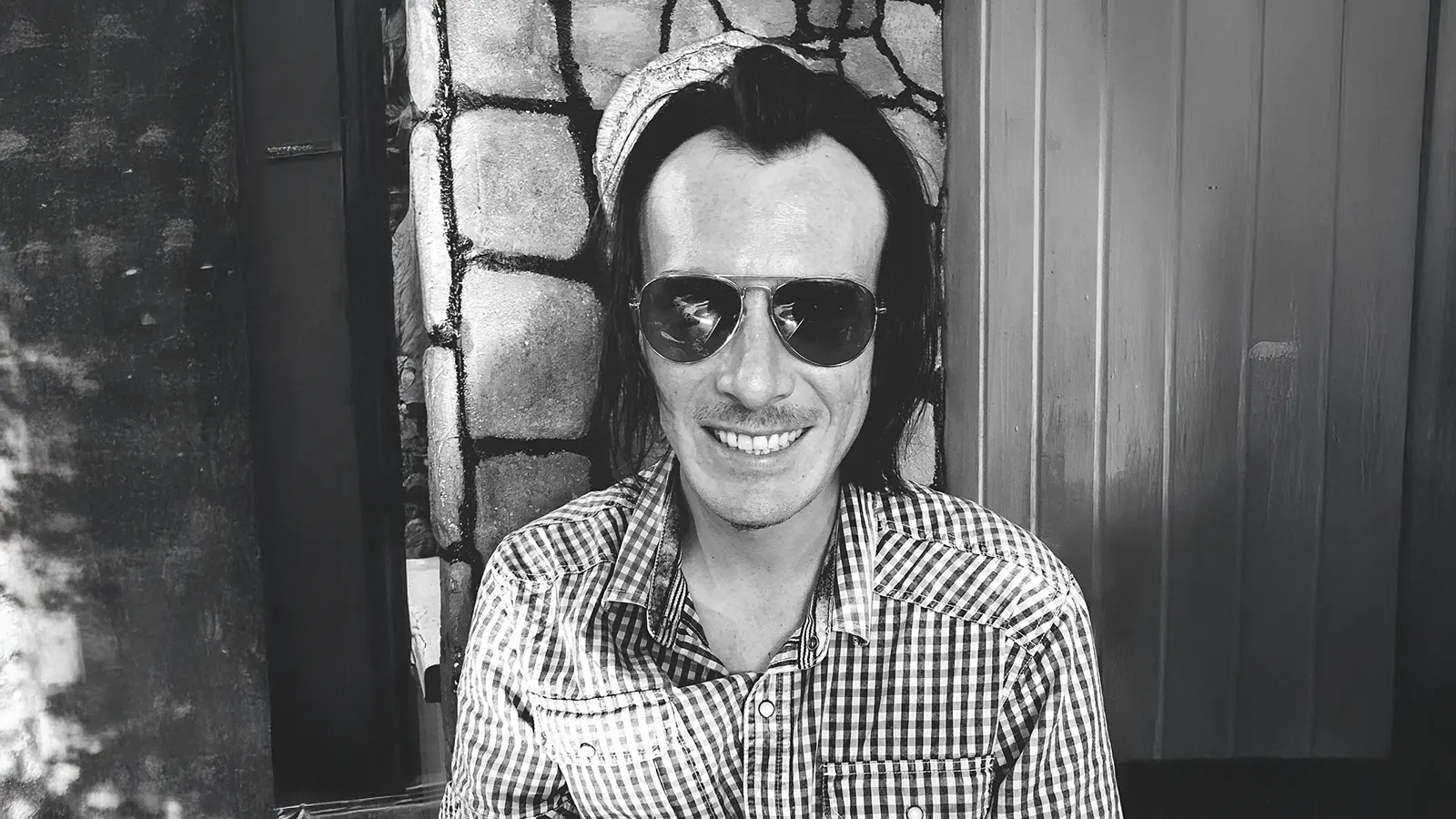 This black and white image features a smiling man in a plaid shirt and aviator sunglasses. Classic aviators have a long history and are representative of the 1950s eyewear trend.