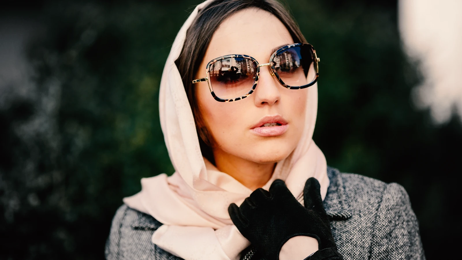 This portrait shows a woman in a grey coat and a light pink scarf wrapped around her head. She is wearing very large, square-shaped designer sunglasses with thick, tortoiseshell and gold frames, exuding a sophisticated and glamorous style.